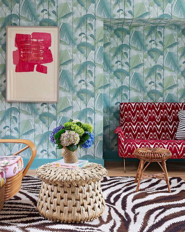 Eclectic living room features a round woven coffee table on a black and white zebra rug, a red chevron sofa with black and white stripe pillow, a rattan stool and teal palm jungle wallpaper.