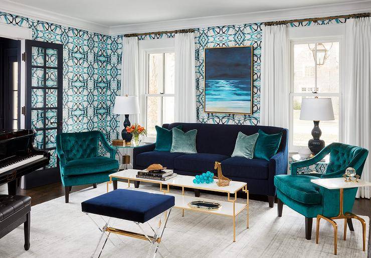 Jewel-toned room colors transform an ordinary room into a lovely eye-catching dwelling space. A long white and brass coffee table is centered atop alight gray rug between two teal velvet tufted accent chairs, a blue lucite stools and a blue velvet sofa with teal accent pillows. Blue and aqua wallpaper dress the walls neutralized by white window curtains and a black glass door panel.