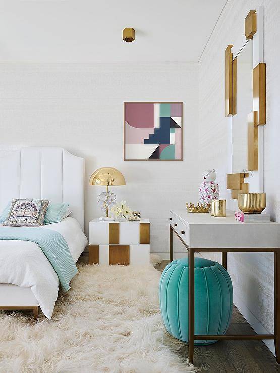 Bedroom features a white channel tufted bed with teal and white bedding atop a cream flokati rug, a white and gold nightstand lit by a gold lamp and a gold and white art deco mirror mounted over a white and gold desk with teal velvet stool.