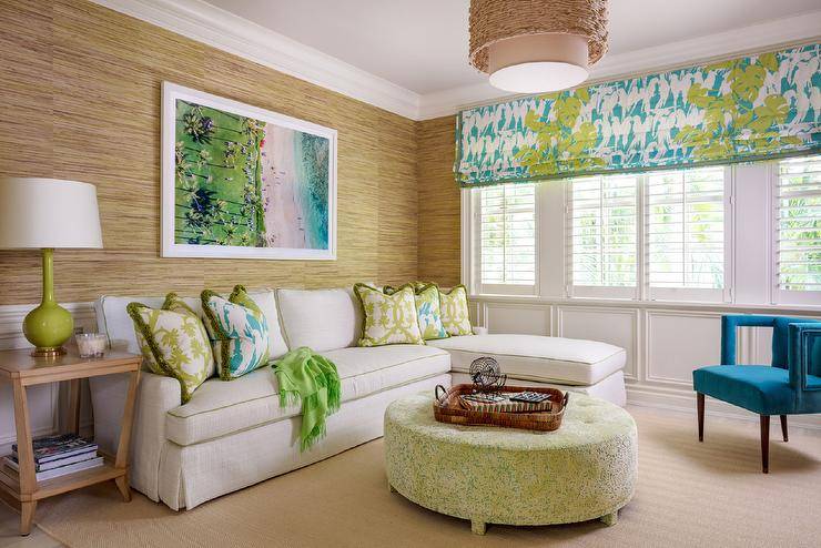 Art hangs from a gold raffia wallpapered wall lined with wainscoting and is positioned over a white sofa trimmed with green piping complementing lime green and teal blue pillows. A lime green lamp sits on a tan end table, while a round yellow and green ottoman is placed in front of the sofa. A chic teal blue velvet chair sits beneath windows fixed above wainscot trim and covered in plantation shutters layered under a teal blue and green roman shade.