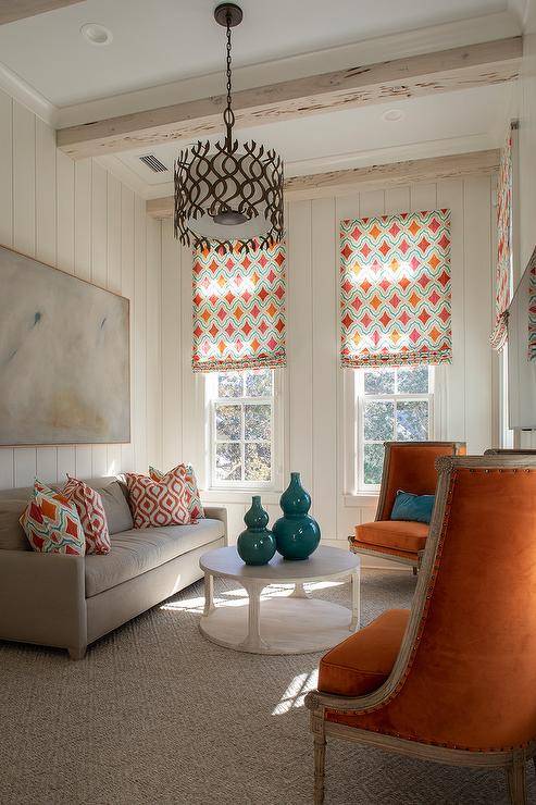 Red trellis pillows on a gray sofa in a small living room paired with a round light wood coffee table and orange velvet French chairs. Ivory shiplap walls add depth and charm to the cottage living space.