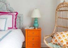 Blue and orange girl's bedroom features a curved headboard upholstered in Brunschwig & Fils Les Touches Fabric on bed dressed in pink and blue bedding placed next to an orange nightstand and a teal triple gourd lamp as well as a Two's Company Hanging Rattan Chair lined with orange pillows.