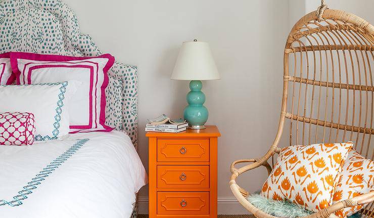 Blue and orange girl's bedroom features a curved headboard upholstered in Brunschwig & Fils Les Touches Fabric on bed dressed in pink and blue bedding placed next to an orange nightstand and a teal triple gourd lamp as well as a Two's Company Hanging Rattan Chair lined with orange pillows.