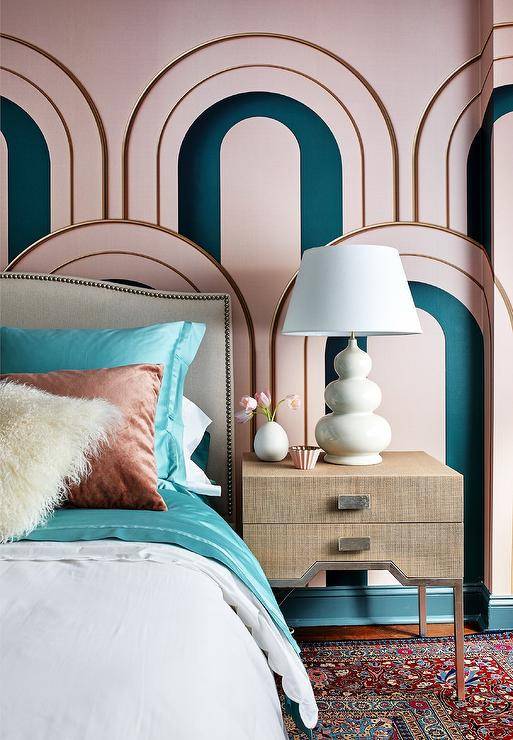 Stylish pink and teal bedroom boasts pink and teal walls complemented with gold trim. A taupe nightstand lit by a gray triple gourd lamp sits beside a gray French linen headboard lined with brass nailhead trim. The headboard finishes a bed dressed in teal bedding topped with a brown sheepskin pillow layered in front of brown velvet pillows.