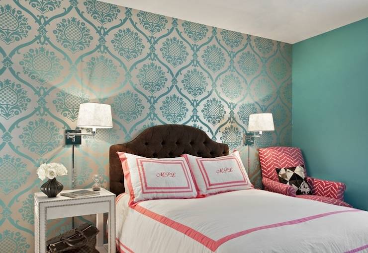 Gorgeous girl's room features accent wall clad in teal damask wallpaper and the rest of the walls painted teal. Chocolate brown tufted velvet headboard on queen bed is dressed in white and pink, monogrammed bedding flanked by polished nickel, swing-arm sconces over single nightstand, white studded nightstand, to the left and pink chevron chair to the right.