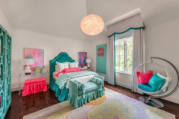 Colorful girl's room features vaulted ceiling accented with feather chandelier over teal velvet headboard on full bed dressed in white and teal bedding, teal monogrammed shams, white and teal chevron duvet, hot pink bolster pillow and teal ruffled bedskirt flanked by hot pink ruffled benches tucked under matching lucite console tables used as nightstands topped with ivory gourd lamps under colorful abstract art alongside turquoise ruffled bench adorned with white, gray and teal striped bolster pillows placed at the foot of the bed atop pastel colored rug. An acrylic swivel bubble chair with hot pink and teal pillows stand in front of window dressed in white scalloped valance with teal trim paired with white curtains with teal ribbon trim.