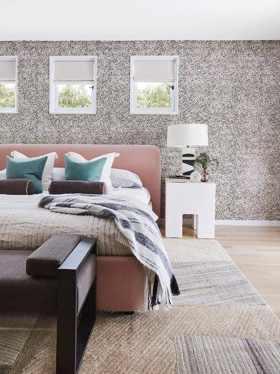 Contemporary bedroom features a blush pink velvet bed accented with teal pillows and black bolster pillows, a chunky white nightstand lit by a black and white block lamp, animal print wallpaper a black bench at the foot of the bed over a colorful rug.