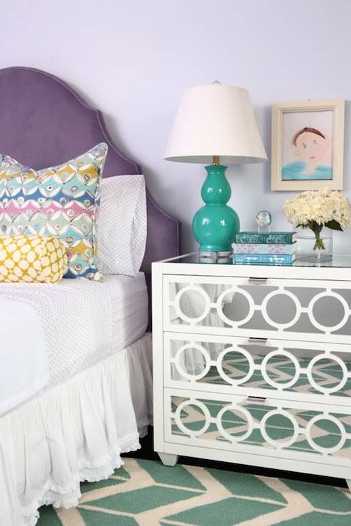 Pretty girl's room with arched purple headboard layered with white bed linens topped with pastel colored pillows situated next to a Worlds Away Ava Nightstand topped with a teal double gourd lamp with framed kid's art above over hardwood floors layered with a teal chevron rug, Jill Rosenwald by Surya Fallon Arrows Teal Hand Woven Flatweave Rug.