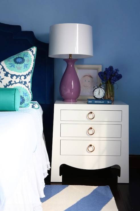 Gorgeous girl's bedroom with navy velvet headboard layered with white bedding topped with teal and blue suzani pillows and a teal bolster pillow flanked by a Bungalow 5 Jacqui 3 Drawer Side Table topped with framed kid's art and a purple ceramic lamp over dark hardwood floors layered with a blue and white striped rug.