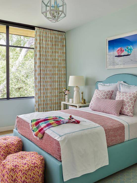 Two orange and red chevron stools sit at the foot of a turquoise blue bed dressed in red bedding topped with layered red pillows. A Gray Malin Two Llamas with Pink and Teal Balloons is framed and hung over the bed, while white bedside tables are styled and lit with Arteriors White Tassel Lamps. Large windows are covered in white and yellow curtains.