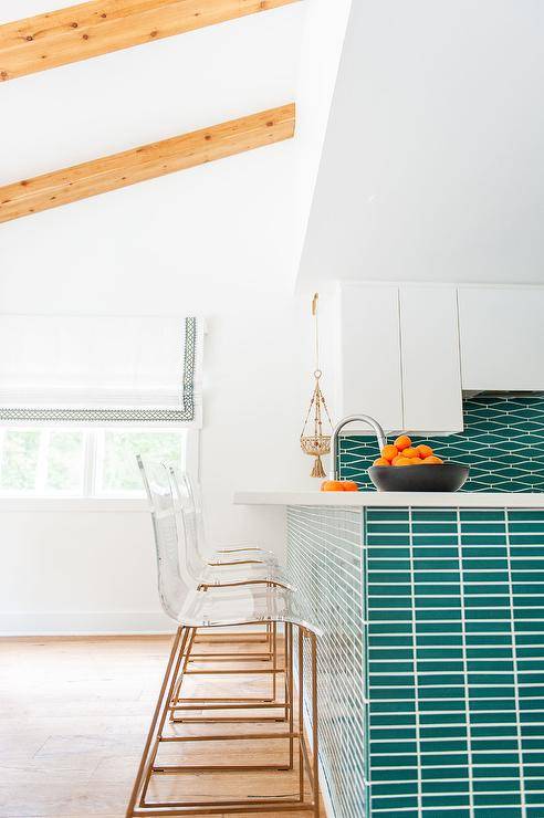 Clear acrylic counter stools sit at a teal tiled kitchen peninsula complementing teal geometric backsplash tiles in a beautifully designed contemporary white and teal kitchen.