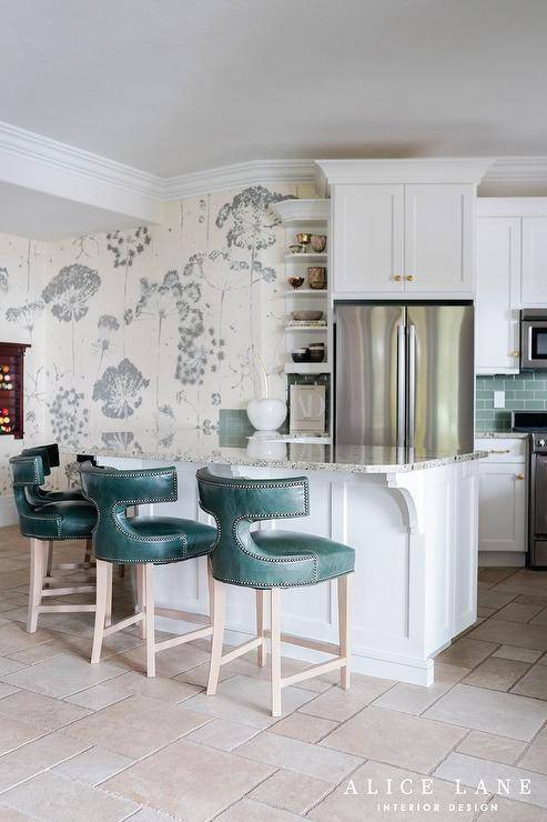 Teal leather t-back stools are placed at a granite countertop accenting a white kitchen peninsula wrapping around to built-in display shelves. A stainless steel double door refrigerator is recessed under white cabinets with brass knobs.