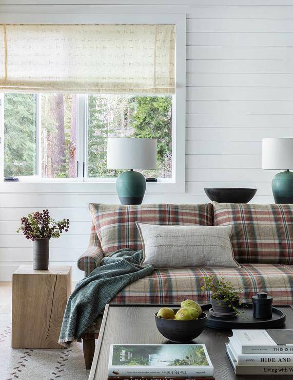 Cottage style living room features a red and gray plaid sofa lit by teal green and black lamps, an oak block end table, a dark brown wooden coffee table and white shiplap walls.
