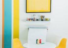 Yellow, teal, and orange stripes accent a white wall holding a yellow print above a white shelf fixed above a wall mount paper holder, as mini yellow molded plastic chairs flank a round white play table with wooden dowel legs.