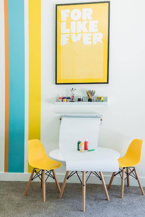 Yellow, teal, and orange stripes accent a white wall holding a yellow print above a white shelf fixed above a wall mount paper holder, as mini yellow molded plastic chairs flank a round white play table with wooden dowel legs.