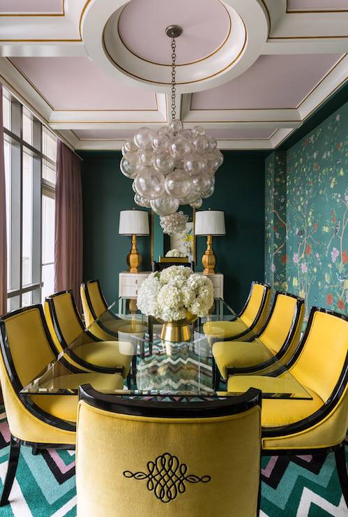 Chinoiserie dining room features a coffered ceiling with coffers painted violet, Sherwin Williams Wallflower, accented with an Oly Studio Muriel Chandelier illuminating an acrylic dining table, Plexi-Craft Elongated X Base Dining Table, lined with yellow dining chairs from Lee Industries atop a teal chevron rug. The back wall of the dining room boasts a teal wall painted Sherwin Williams Rookwood Sash Green lined with a white sideboard cabinet topped with gold buffet lamps, Shine by Sho Empire Table Lamps. A wall of windows is dressed in violet curtains facing an accent wall clad in de Gournay Earlham Wallpaper.