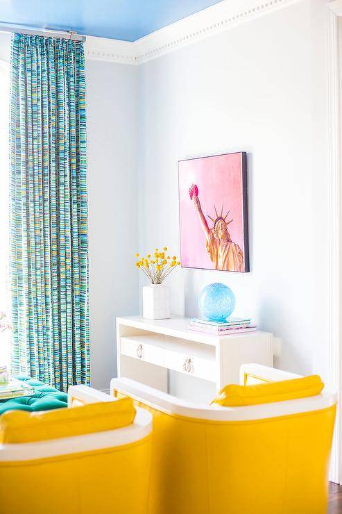 Contemporary living room furnished with a set of canary yellow accent chairs facing a teal tufted ottoman. Colors make bold statements in every corner of this space- a white lacquer console table features styled decor with a pink statue of liberty wall art overhead. Dentil crown moldings accent the room along with a bright blue ceiling complimenting blue and green colorful curtains creating an aesthetically pleasing finish.