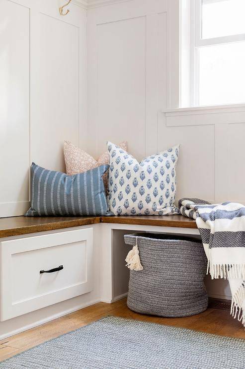 Mudroom features whiteboard and batten trim, brown wood built-in bench, white storage drawers, black pulls, blue and white striped throw, blue and white accent pillows, and gray woven basket. with gray rugs.