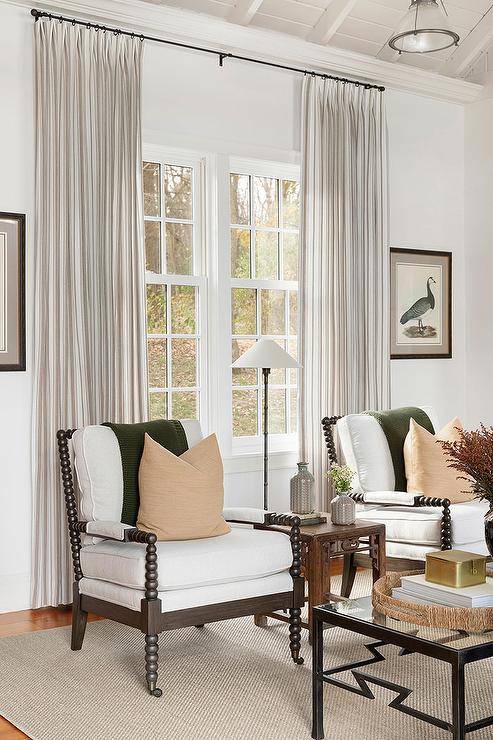 The cottage living room boasts a small wooden side table in front of a window with long pleated cream curtains and a black spindle chair with a forest green throw blanket.