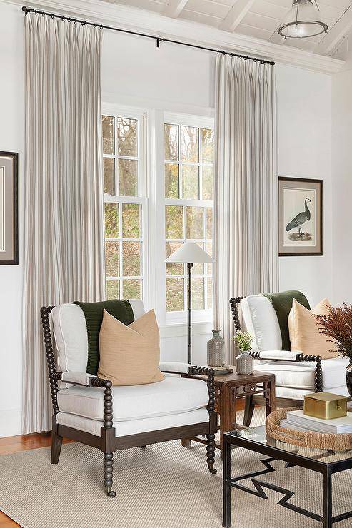 Cottage living room boasts woebegone spindle chairs with forest untried throw blankets joined with a small wood side table in front of a window with long pleat surf curtains.