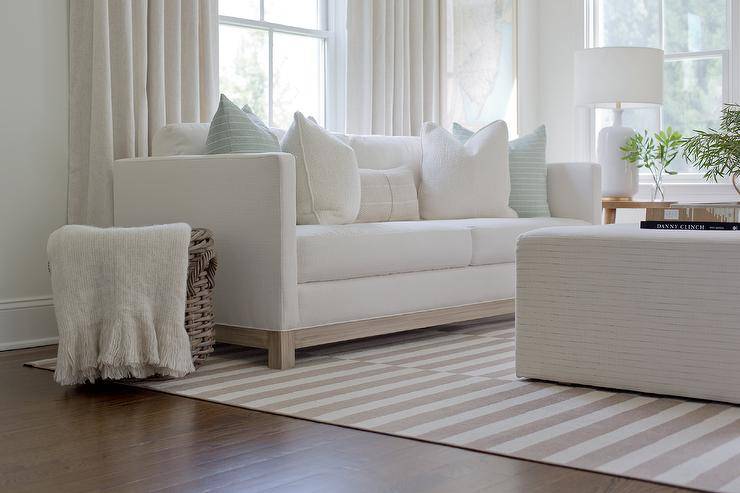 Living room features a white shelter back couch with light blue stripe pillows with an end table lit by a white lamp, a white ottoman as a coffee table over a light gray striped rug and a cream throw blanket on a woven basket