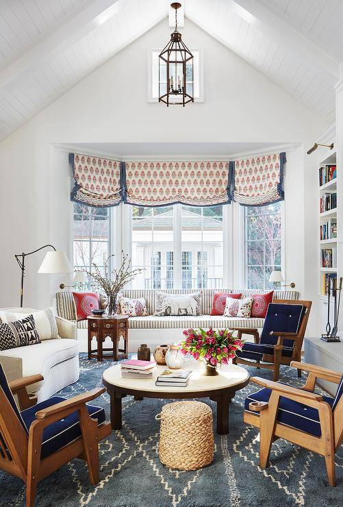 A Moroccan accent table sits on a blue rug in front of a built-in bay window bench topped with gray striped cushions accented with red pillows. The windows are covered in red and blue roman shades.