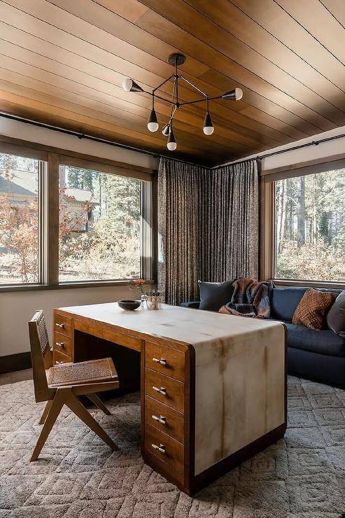 An oil rubbed bronze atom light fixed to a brown plank ceiling illuminates a vintage desk paired with a cane chair placed on a beige diamond print rug in front of a blue sofa located beneath a window.