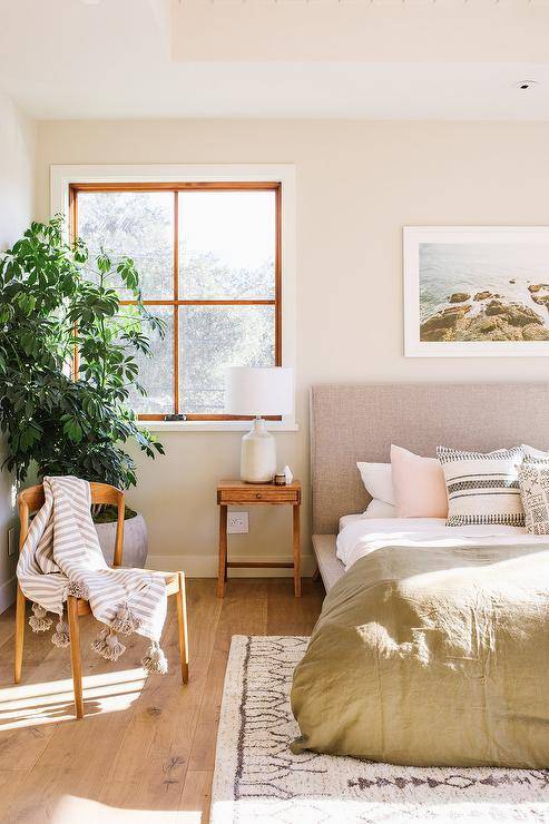 Blush pink pillow on a gray platform bed in a transitional bedroom with warm natural light. The heather gray platform bed is dressed with an olive green duvet color beside a vintage wood nightstand displaying a white lamp. A Scandinavian style wood chair displays a pompom throw blanket for a comfy and stylish finish.