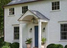 A beautiful white home boasting white siding and a blue front door positioned beneath a portico lit by an oil rubbed bronze carriage sconce.