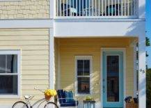 Side view of a yellow two story house with a turquoise blue front door under a second level balcony. A turquoise blue bench decorates the outdoor porch along with a blue and wood top side table for a cozier and personalized space.