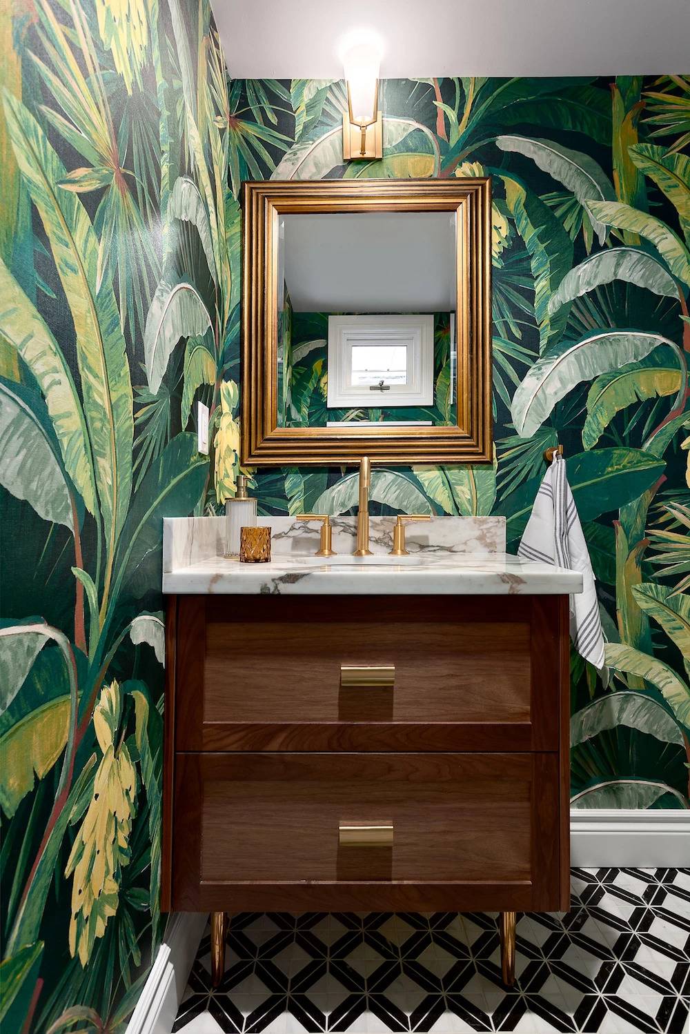 bathroom with bright green tropical leaf wallpaper, small dark wood vanity and gold mirror