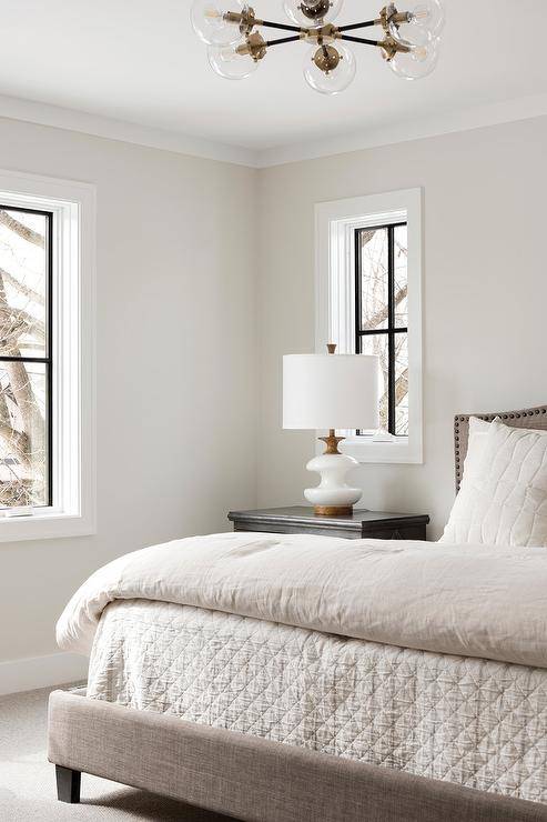 Transitional bedroom boasts a beige linen camelback bed with white and beige bedding beside a black nightstand displaying a white and gold lamp. Simple neutrals in soft and soothing hues making the bedroom look timeless and sophisticated finished with a glass globe chandelier contemporary chandelier.