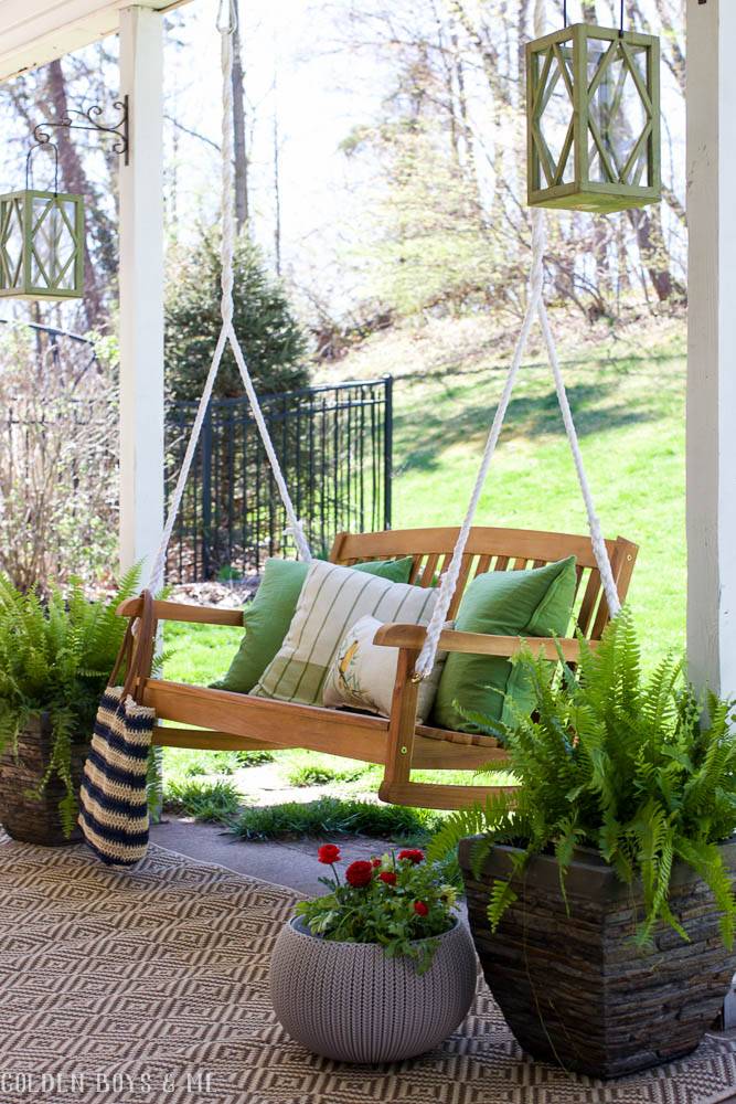 wooden patio swing chair with bright green pillows, patterned rug and green potted plants