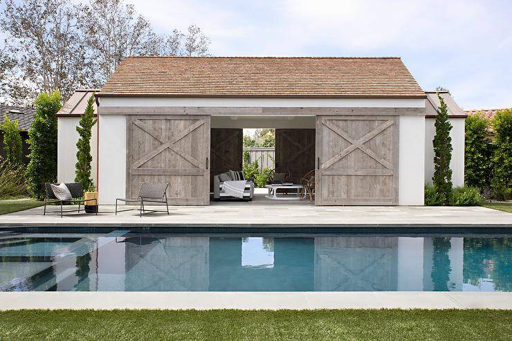 An in ground swimming pool is located in front of a white pool house accented with gray wash dual barn doors on rails.
