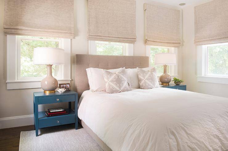 A series of bedroom windows dressed with beige roman shades surround a bed with a beige tufted headboard and white bedding. Beige and white accent pillows with boho pattern compliment the neutral theme while blue nightstands flank the bed displaying beige gourd lamps topped with white drum shades.