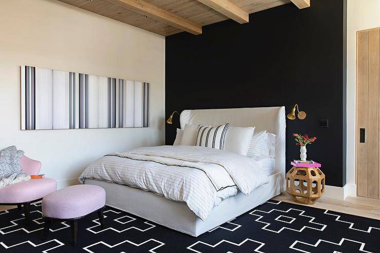 Chic black and white bedroom is styled with a white wingback bed dressed in gray striped bedding and topped with an ivory and black pillow. The bed sits on a black geometric rug beside a carved wood bedside table lit by a brass sconce fixed to a black accent wall. A long black and white abstract art piece hangs from a white wall to the side of a pink chair and ottoman.