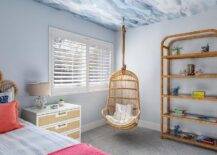 A rattan hanging chair hangs from a blue wallpapered ceiling in the corner of a bedroom between a vintage rattan etagere and a window covered in plantation shutters. A white nightstand with tan fabric drawers is lit by a coral lamp and placed in front of a light blue wall.
