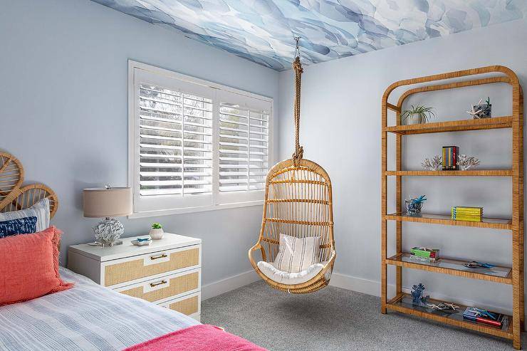 A rattan hanging chair hangs from a blue wallpapered ceiling in the corner of a bedroom between a vintage rattan etagere and a window covered in plantation shutters. A white nightstand with tan fabric drawers is lit by a coral lamp and placed in front of a light blue wall.