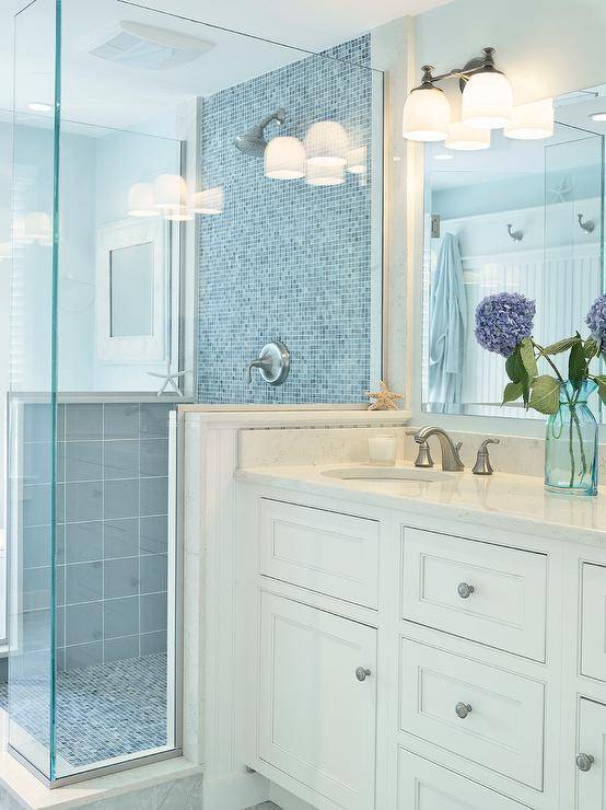 Beachy bathroom features a large walk-in shower centered against the vanity wall boasting sky blue tiled sides and a blue glass mosaic tile wall and shower floor beside an ivory shaker vanity adorned with brushed nickel pulls alongside a white quartz counter finished with a beveled vanity mirror above lit by a frosted glass vanity light.