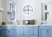 Beautiful coastal bathroom with built-in sky blue dual sink vanity accented with nickel pulls alongside white counters which frame oval porcelain sinks situated below white vanity mirrors on tongue and groove clad walls lit by white enamel pendants on either side of a round window. The blue double vanity stands atop an inset wood trellis patterned floors framing marble tile laid on the diagonal.