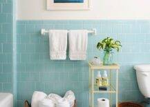 Pretty bathroom with aqua blue tiled half walls and bath surround. The blue from the tile is picked up in the coastal print hanging on the ivory painted wall above. Mosaic tiles in varying shades of blue cover the floor and add to the watery color palette. A large seagrass basket holds rolled towels whilst a pale yellow metal and glass etagere holds flowers and other bath essentials.