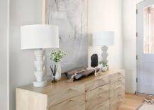 A light gray abstract art piece hangs in an elegant foyer beneath a gray plank ceiling and over a light brown credenza topped with white spindle lamps.