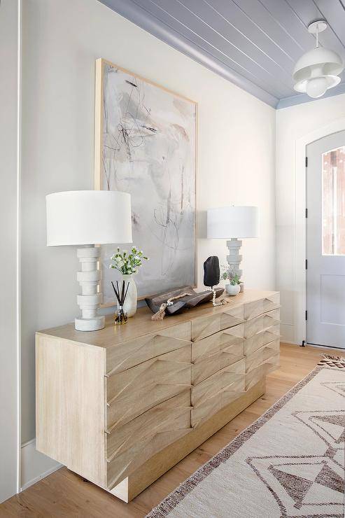 A light gray abstract art piece hangs in an elegant foyer beneath a gray plank ceiling and over a light brown credenza topped with white spindle lamps.