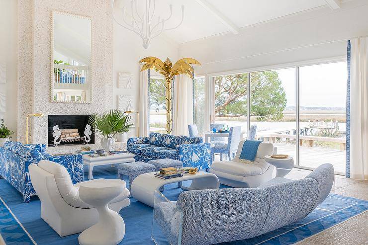 Blue palm leaf sofas and blue chevron pattern stools join with a white chinoiserie coffee table in a living room finished with a mosaic stone fireplace and a white spiky mirror.