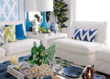 Blue and green tropical style living room with panels of turquoise blue Moroccan tile stencil beside turquoise framed mirror panels which highlight a modern white side table topped with turquoise bottle shaped lamp beside a white armless sofa layered with blue and green chevron pillows and a blue velvet pillow. A Fiddle Leaf Fig stands in the far corner, next to sun filled windows dressed in floor length sheers, with a pair of armless white accent chairs across from a navy tufted ottoman coffee table laden with books and a silver bar tray with leather handles.
