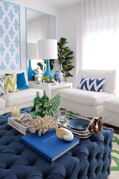 Blue and green tropical style living room with panels of turquoise blue Moroccan tile stencil beside turquoise framed mirror panels which highlight a modern white side table topped with turquoise bottle shaped lamp beside a white armless sofa layered with blue and green chevron pillows and a blue velvet pillow. A Fiddle Leaf Fig stands in the far corner, next to sun filled windows dressed in floor length sheers, with a pair of armless white accent chairs across from a navy tufted ottoman coffee table laden with books and a silver bar tray with leather handles.