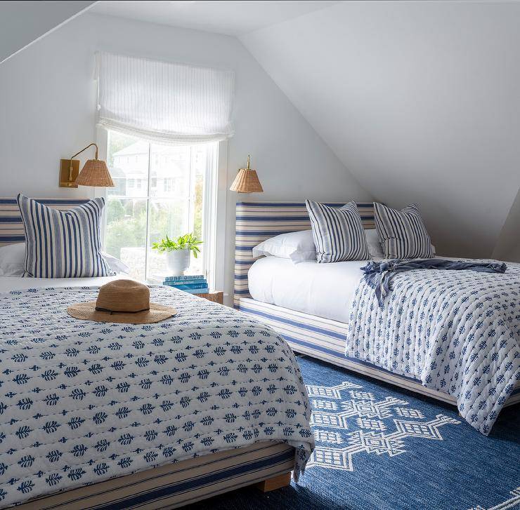 A sloped attic ceiling is fixed over a shared white and blue cottage bedroom featuring cream and blue striped beds topped with blue striped pillows placed on white and blue bedding. The beds, placed on a blue rug, are lit by brass and rattan sconces and flank a window covered in a white roman shade.