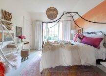 This stylish, boho chic kid's bedroom boasts an orange ombre accent wall positioned behind an iron canopy bed. The bed sits on a pink a pink and orange vintage wool rug and is accented with pink and purple pillows, while a white hanging chair is fixed in a corner.