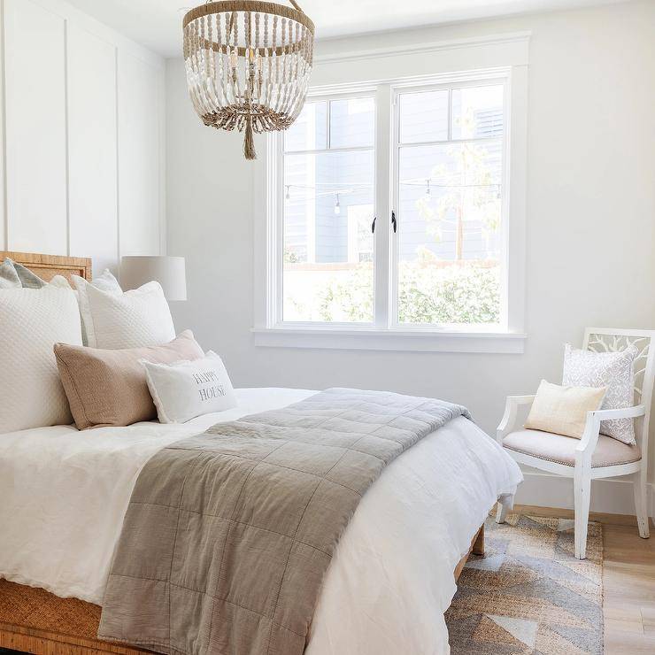 Restful cottage bedroom is illuminated by a white and tan beaded chandelier hung over a Serena & Lily Balboa Bed placed against a board and batten wall and accented with pink and gray bedding. The bed is placed on a tan and gray rug, while a white wooden chair with a light gray cushion is positioned in a corner beside uncovered windows.