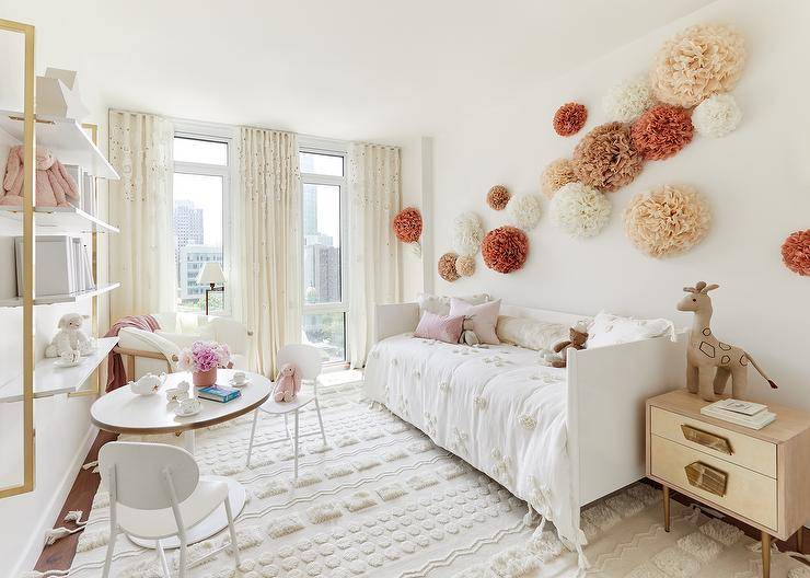 Paper pom poms are mounted over a white daybed dressed in embroidered flower bedding and placed on a Moroccan wedding blanket rug beside a gold nightstand. Ivory pleated curtains cover windows located on a wall adjacent to white and gold wall mounted shelves fixed over a modern play table and chairs.
